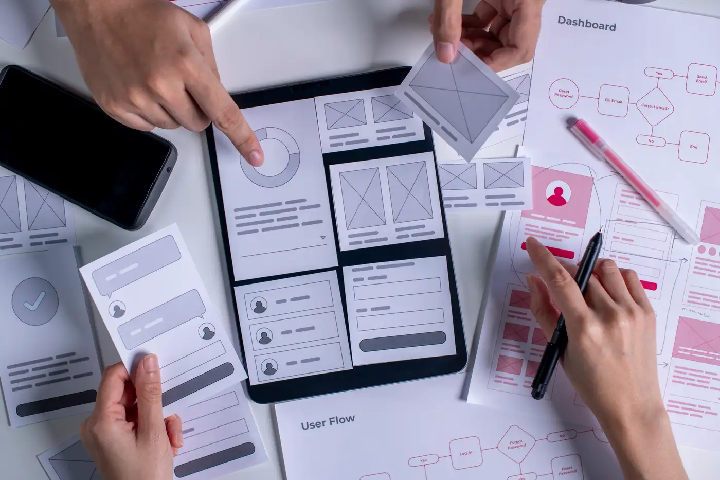 Designers sorting paper wireframes and user-flow sketches around a tablet on a desk