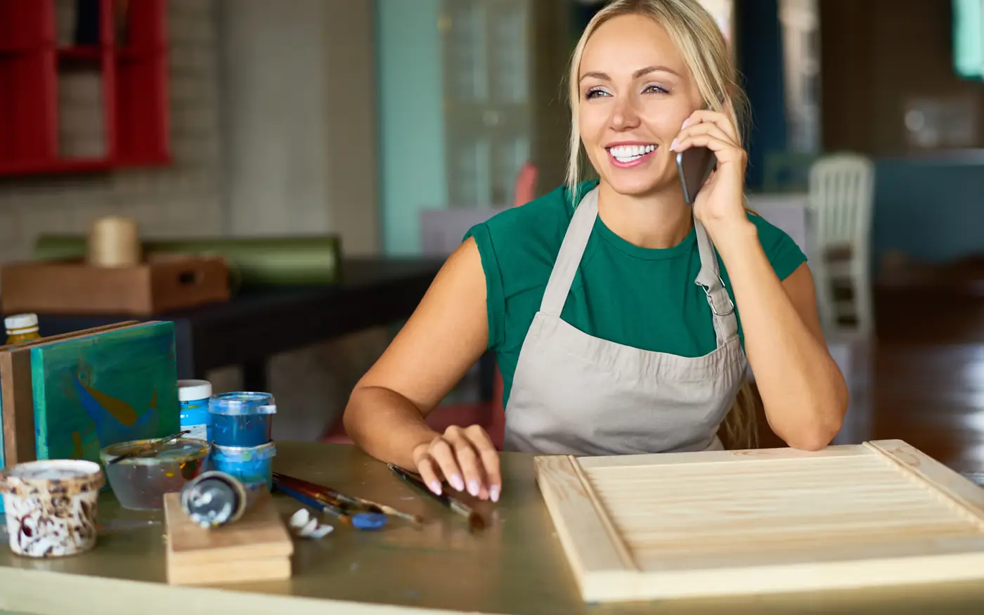 Small business owner in an apron smiling while she takes a customer call on her phone at her workbench