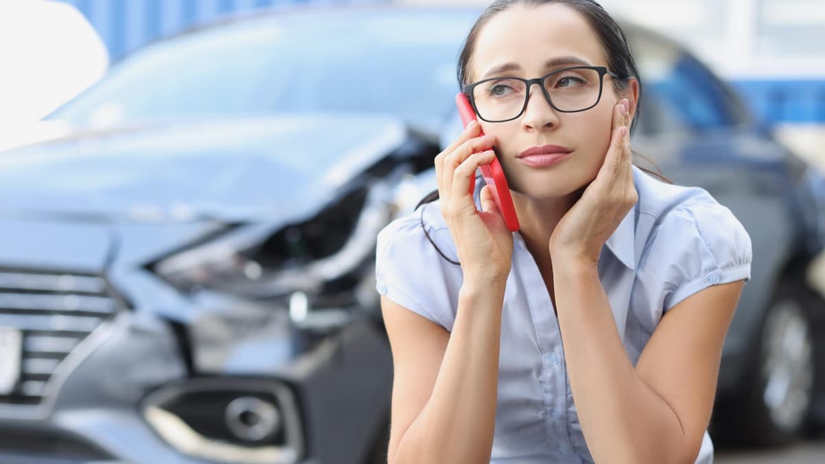 A woman in glasses talks on her phone, hand to her head, beside a car with a crushed front end