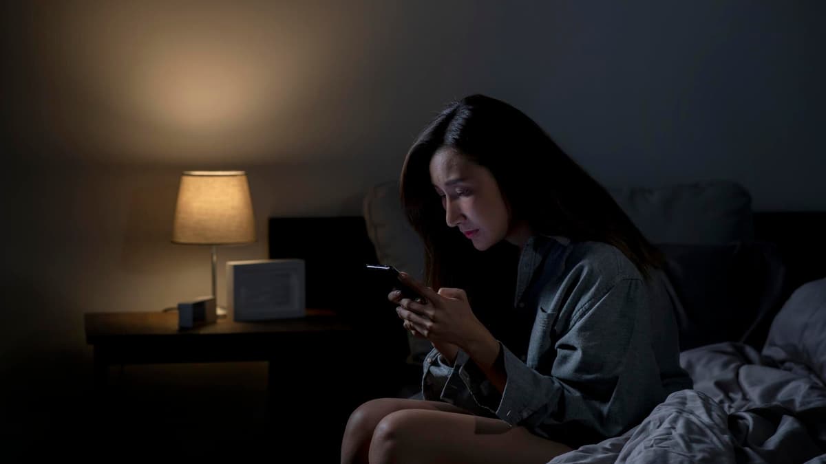 A young woman sits on the edge of her bed at night, lit by a lamp, typing on her phone