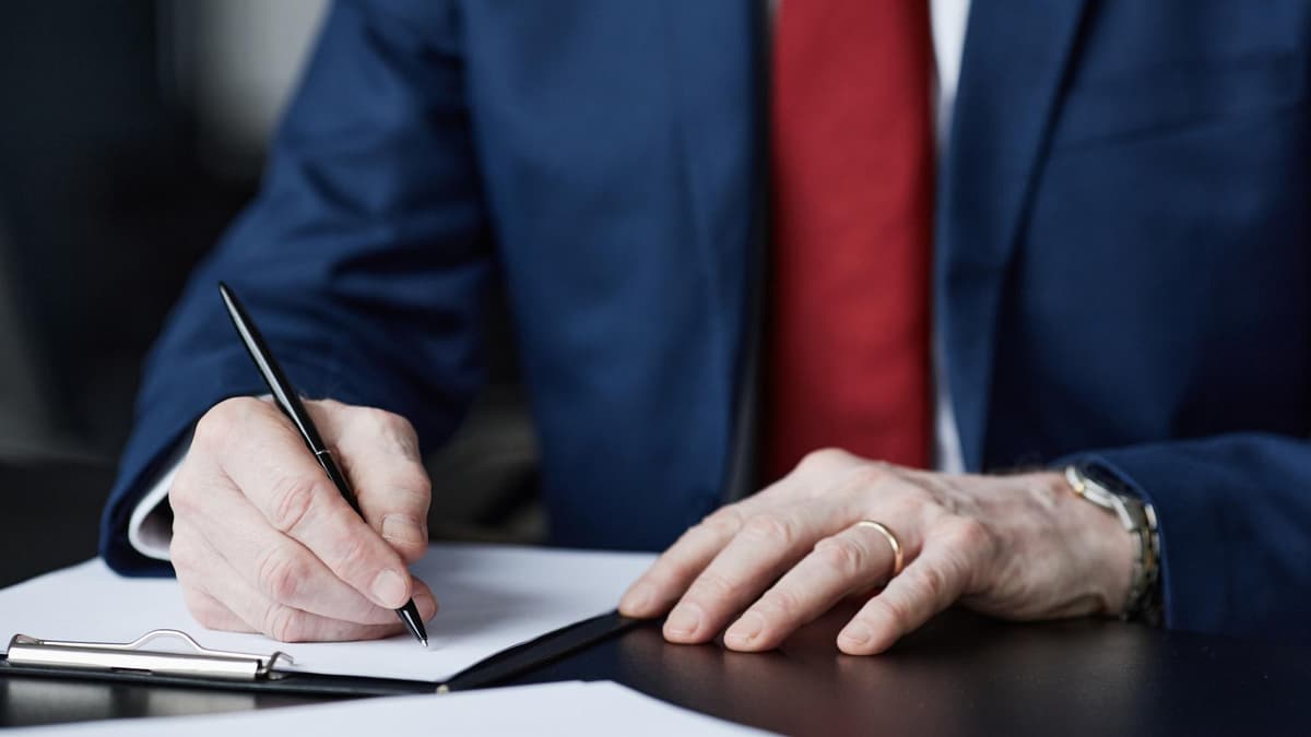 A man in a navy suit signs a contract at an office desk