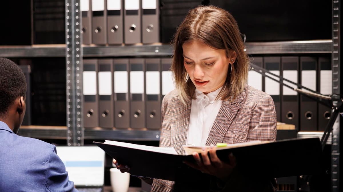 A woman in a checked blazer reads an open case binder in a file room lined with shelves