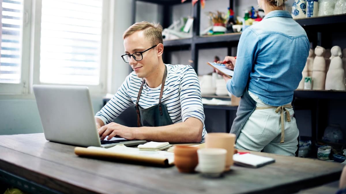 Small shop owner updating his business website on a laptop while his partner checks orders