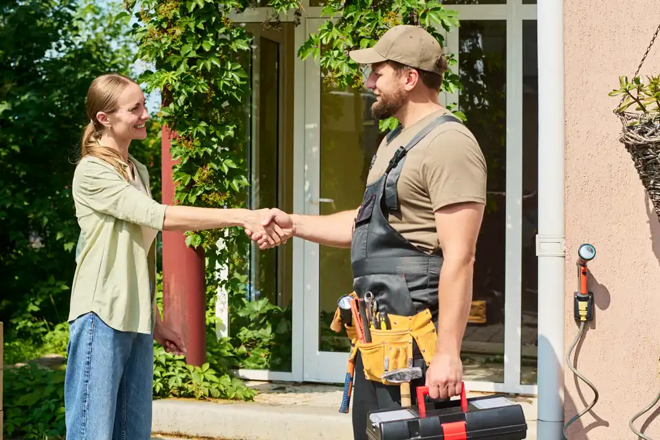 A homeowner shakes hands with a tradesman carrying a toolbox outside her front door