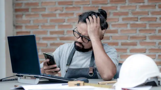 A tradesman at his desk holds his head while checking his phone