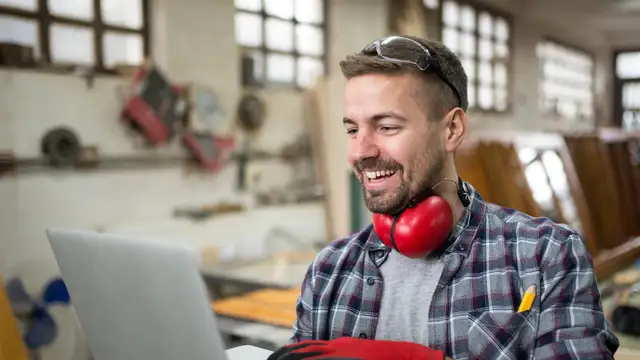 A carpenter smiles at his laptop in his workshop